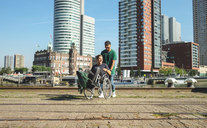 Een jongen duwt een andere jongen in een rolstoel over een plein in een stad. 