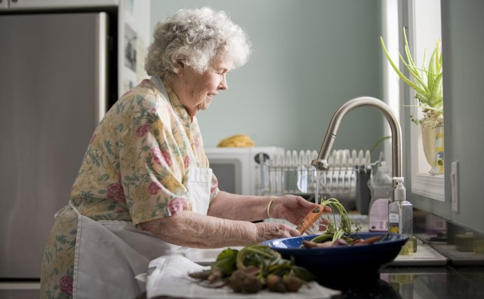 Oudere vrouw in de keuken wast groenten af onder de kraan.