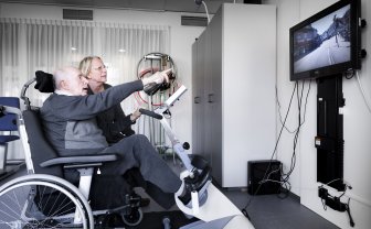 man zit in een rolstoel en fietst met zijn benen terwijl hij naar een scherm kijkt met een weg daarop.
