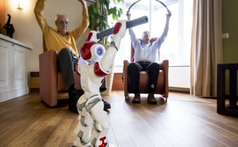 robot Zora doet oefening voor, twee ouderen op achtergrond doen het na