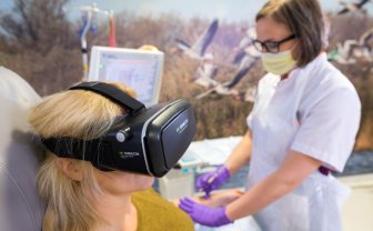 Vrouw met virtual reality bril op wordt in haar arm geprikt door een verpleegkundige.