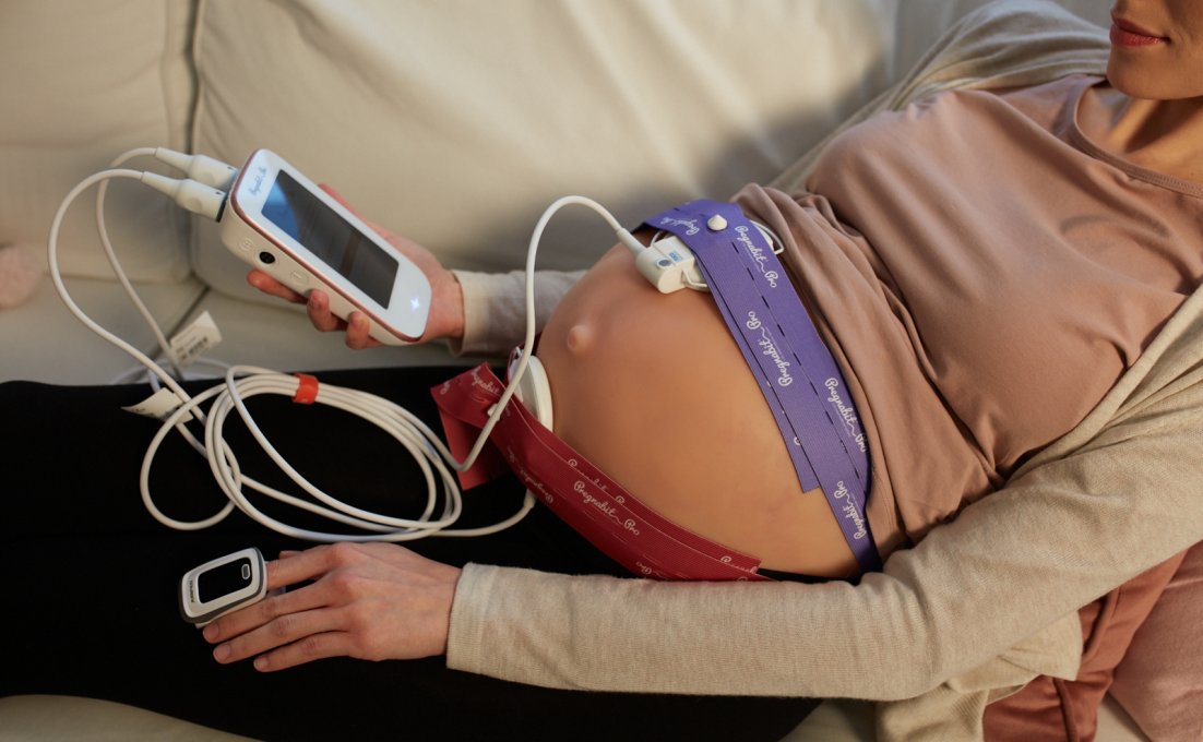 Een zwangere vrouw ligt op de bank en heeft het PregnaOne-apparaat aangesloten op haar buik