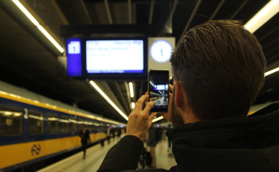 Een man op een perron richt de camera van zijn smartphone op een bord met reisinformatie van de NS.  