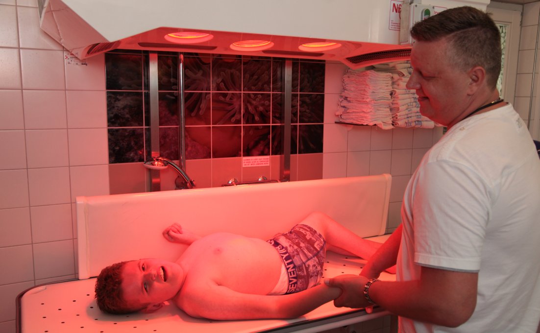 Een jongen met een lichamelijke beperking ligt op tafel onder infraroodlampen. Een man houdt zijn hand vast.