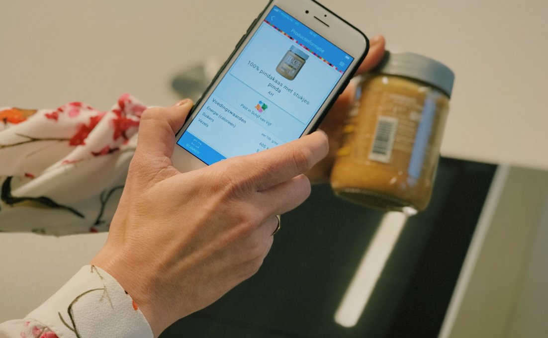 Een mevrouw scant een pot pindakaas met de app Kies ik gezond?. Op het scherm verschijnt informatie over het product