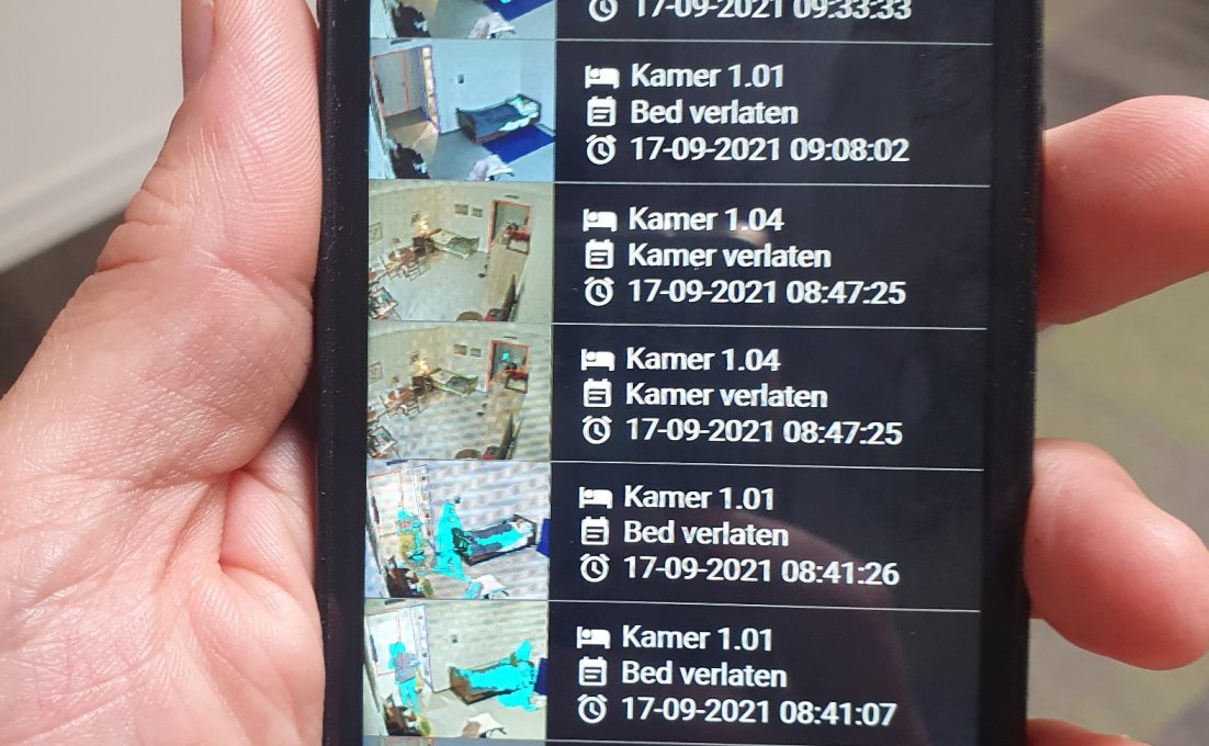 Op een telefoon is de app van SmarterCare geopend. Daarop worden verschillende kamers van patiënten weergegeven. 